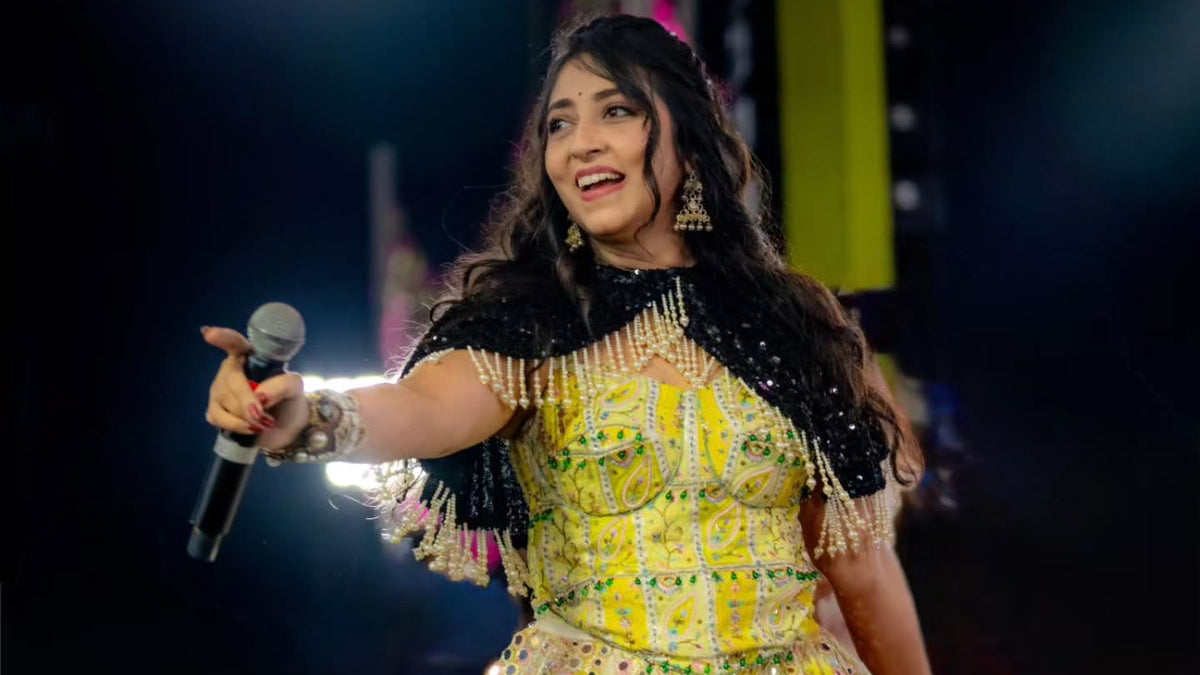 A woman performing on stage wearing a bright yellow and white embellished traditional dress with black accents, holding a microphone. The background is dark with stage lighting.